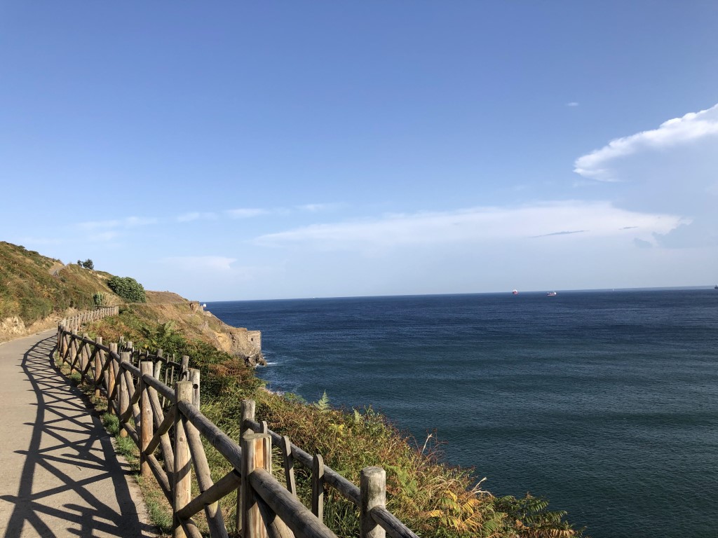 Blå himmel med havet till höger. Promenadstig med staket mot kusten.