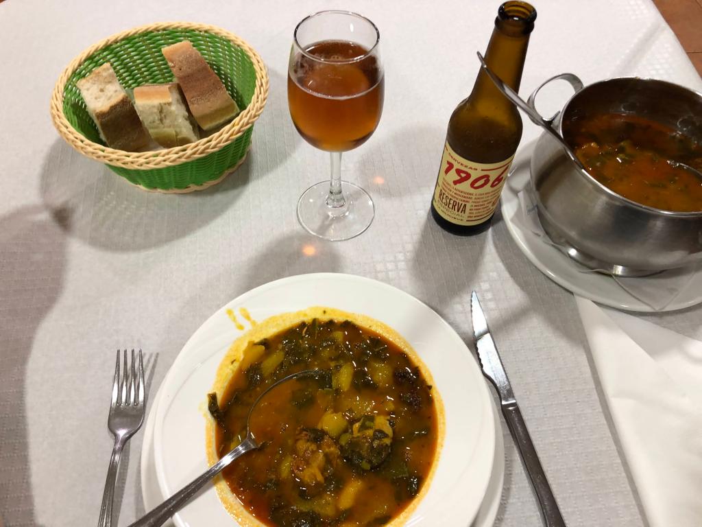 A plate full of soup with potatoes and sausages, a glass of beer and some bread.