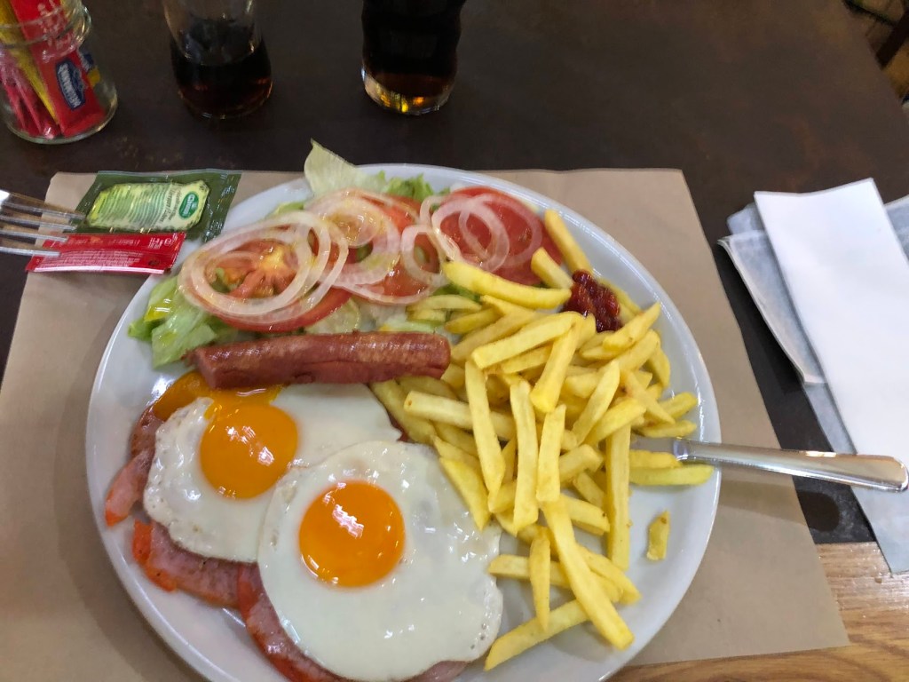A plate with two fried eggs on ham, a sausage, salad and fries. Coke in a glass.